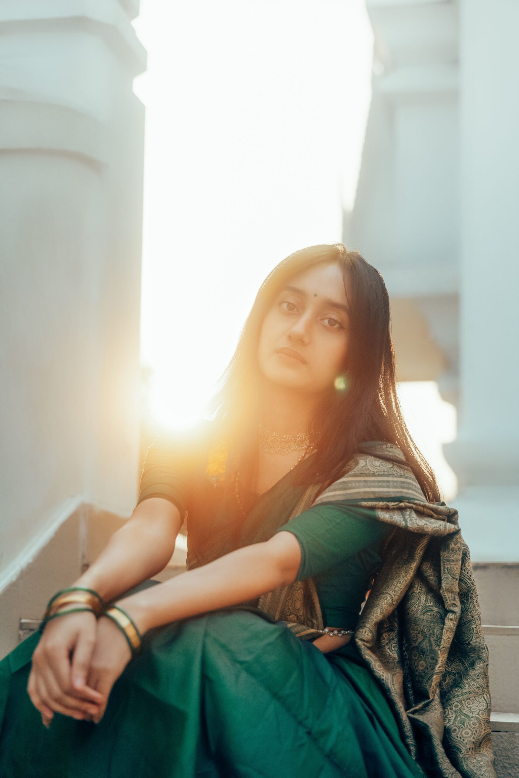 A young woman in a green saree sits gracefully on stairs, radiating elegance in the warm afternoon sunlight.